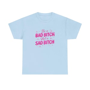 May include: Light blue t-shirt with the text "Be a BAD BITCH Not a SAD BITCH" in pink. The text is surrounded by small star-like shapes. The shirt has a crew neck and short sleeves.