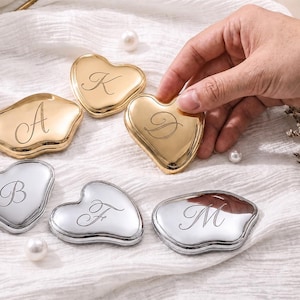 Peut inclure: Une collection de six miroirs de poche en métal en forme de cœur avec des finitions dorées et argentées polies. Chaque miroir présente une lettre cursive gravée, incluant A, K, D, B, F et M. Les miroirs sont disposés sur un tissu blanc.