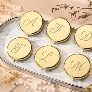 May include: Six round gold-toned compact mirrors with polished metallic surfaces. Each mirror features a single engraved letter in a cursive script, specifically the letters A, F, D, T, L, and M, centered on the front of the circular casing.