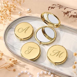 May include: Four round gold-toned compact mirrors with polished metallic finishes. Three mirrors are closed, each engraved with a single cursive letter: A, T, or M. One mirror is open, revealing two reflective surfaces inside the circular gold frame.