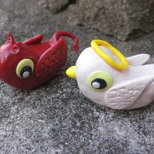 Puede incluir: Dos pequeñas figuritas de cerámica, una roja con cola y cuernos, la otra blanca con un halo amarillo. Ambas tienen ojos negros y picos amarillos.
