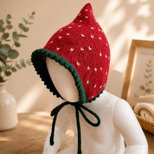 Puede incluir: Un gorro de punto rojo con la parte superior puntiaguda, con pequeños detalles bordados en amarillo que parecen semillas de fresa. El gorro tiene un borde acanalado de color verde oscuro alrededor de la cara y dos lazos de punto largos.