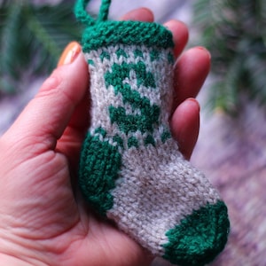 Puede incluir: Un pequeño calcetín de Navidad tejido a mano, en verde y gris. El calcetín tiene un puño, talón y puntera verdes, con un diseño de patrón verde y blanco en el cuerpo. Una presilla verde en la parte superior.