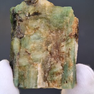 以下が含まれることがあります： 緑、茶色、白の色合いが混ざった、未加工のエメラルド原石。未研磨の鉱物は、目に見える結晶構造を持つテクスチャ加工された表面を持っています。エメラルドは手袋をはめた手で持たれています。