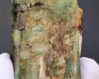 May include: A rough-cut emerald gemstone with a mix of green, brown, and white hues. The raw mineral displays a textured surface with visible crystalline structures. The emerald is held by gloved hands, showcasing its natural, unpolished state.