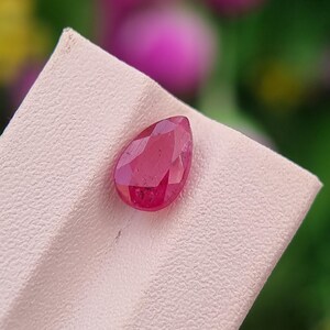 May include: A faceted, pear-shaped ruby gemstone. The gem is a deep, vibrant red color and is displayed on a light pink surface. The background is a soft blur of green, pink, and yellow.