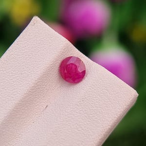 Könnte beinhalten: Ein facettierter, runder, tiefroter Rubin-Edelstein. Der Rubin ist auf einer hellrosa Oberfläche platziert, mit einem verschwommenen Hintergrund aus grünen und rosa Blumen. Der Edelstein ist ein lebendiger, auffälliger Blickfang.