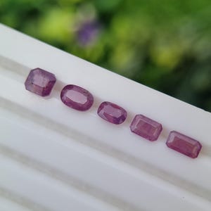 May include: Six purplish-red gemstones in different shapes, including square, oval, and rectangular. The gems are displayed in a row on a white surface, with a blurred green backdrop.