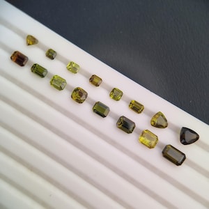 Puede incluir: Una variedad de gemas facetadas en varias formas y tamaños, incluyendo cortes esmeralda, cuadrados y triángulos. Las gemas varían en color desde verde oliva hasta marrón dorado, exhibidas en una bandeja blanca sobre un fondo oscuro.