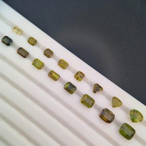 Puede incluir: Una variedad de gemas facetadas en diversas formas y tonos de verde, amarillo y marrón. Las gemas están dispuestas sobre una bandeja blanca con ranuras paralelas, sobre un fondo oscuro. Las formas incluyen rectángulos y cuadrados.