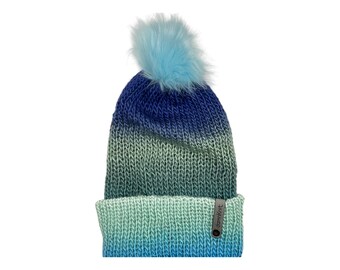 May include: A knitted beanie in shades of blue and green, with a fluffy light blue pom-pom on top. The hat features a folded brim and a small tag that reads "damknit".