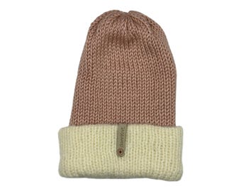 May include: A knitted beanie in a light rose color with a cream-colored folded brim. The hat has a ribbed texture and a small tag with the text "damnKnit" and a button detail.