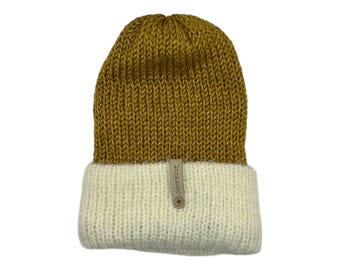 May include: A knitted beanie in mustard yellow and cream. The upper portion is mustard yellow, while the folded brim is cream-colored. A small tag with the text "damnknit" is attached to the brim.