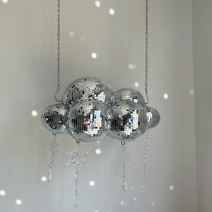 May include: A cloud-shaped disco ball light fixture. The fixture is composed of several mirrored spheres of varying sizes, suspended from chains. Clear snowflake and icicle-shaped crystal ornaments hang below the spheres. The background is a white wall with light reflections.