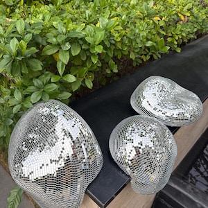 Puede incluir: Tres esculturas abstractas decorativas con superficies espejadas. Las esculturas son de diferentes tamaños y formas, reflejando el entorno. Se colocan sobre una superficie negra con follaje verde en el fondo.