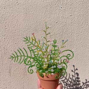 Puede incluir: Un arreglo de plantas en miniatura con cuentas en una pequeña maceta de terracota. La planta presenta hojas y tallos de cuentas verdes, con cuentas de colores que representan flores. La maceta se sostiene contra un fondo marrón claro.
