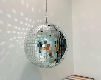 May include: A silver disco ball, composed of small mirrored squares, hangs from a chain. The reflective surface scatters light, creating a pattern on the white wall. The disco ball is a classic party decoration.