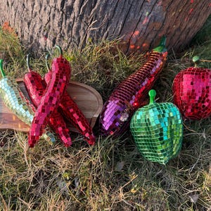 Könnte beinhalten: Eine Sammlung dekorativer Obst- und Gemüseobjekte aus Mosaikglas, darunter rote und grüne Chilischoten, eine lila Aubergine und rote und grüne Äpfel. Die Artikel sind auf einem Holztablett und Gras angeordnet.