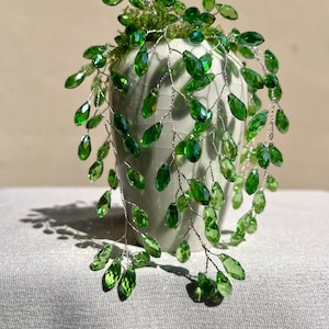 Könnte beinhalten: Eine dekorative Vase mit einem grünen, perlenbesetzten Rankenmuster. Die Vase ist hellgrün und hat eine strukturierte Oberfläche. Die Perlenranke besteht aus grünen, facettierten Perlen und Silberdraht.