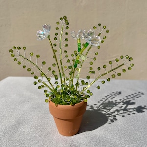 May include: A handcrafted beaded flower arrangement in a small terracotta pot. The arrangement features delicate wire stems with green beads and iridescent flower accents. The pot is approximately 3 inches in diameter.
