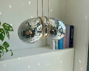 May include: Two silver disco balls, covered in small mirrored squares, are suspended from the ceiling by twine. The disco balls are joined together with twine. The background is a white wall with a shelf and some books.