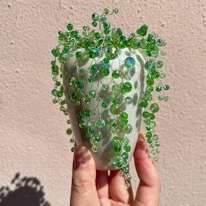 Könnte beinhalten: Eine kleine, weiße Keramikvase mit einem gesprenkelten Muster, verziert mit Kaskaden aus grünen, irisierenden Perlen. Die Perlen sind auf Silberdraht aufgefädelt, wodurch ein zartes, pflanzenähnliches Aussehen entsteht. Die Vase wird in einer Hand gehalten.