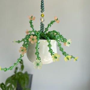 Puede incluir: Decoración colgante de plantas con cuentas. Una maceta de cerámica blanca contiene follaje de cuentas verdes y detalles florales en colores pastel. La planta cuelga de una cadena de cuentas, creando una pieza caprichosa y decorativa.