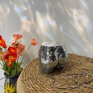 May include: A silver disco ball shaped purse with a chain strap sits on a woven surface. The purse is covered in small, reflective squares. A vase with orange and red flowers is to the left.