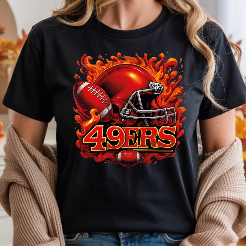 May include: Black t-shirt featuring a vibrant graphic of a red football helmet and football engulfed in flames, with the text "49ERS" in bold red and gold lettering. A football is also at the bottom of the design.