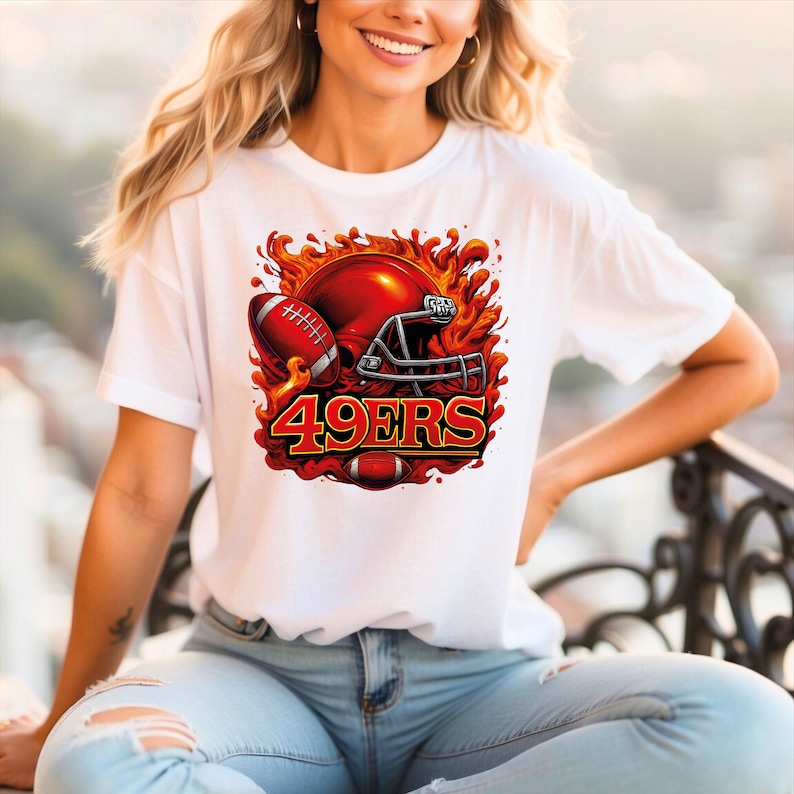 May include: White t-shirt featuring a vibrant graphic of a football helmet, football, and the text "49ERS" in red and gold. The design is surrounded by flames, creating a dynamic and energetic look. Perfect for football fans.