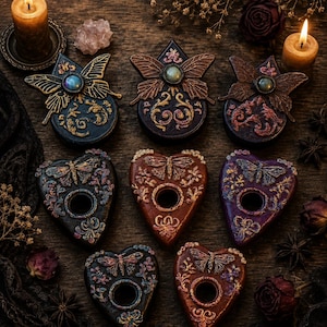 May include: A collection of eight decorative planchette-shaped objects arranged on a dark wooden surface. Three top pieces feature butterfly motifs with central iridescent stones, while five heart-shaped pieces include moth designs and circular cutouts.