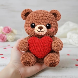 May include: A handmade brown crochet teddy bear holding a red heart. The bear has large black eyes, a tan muzzle, and pink cheeks. The plush toy is a charming gift for Valentine's Day or any special occasion.