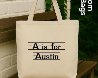 May include: Cream-colored tote bag with black text that reads "A is for Austin" in a school-like font. The bag has two handles and is sitting on a wooden bench. The background includes a white brick wall and greenery.
