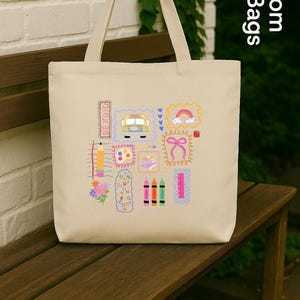 May include: Cream canvas tote bag featuring a colourful school-themed design. The design includes a school bus, rainbow, pencils, and the words "BE KIND". The bag has long handles and the text "Custom Tote Bags US".