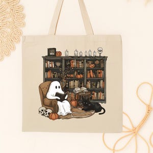 Puede incluir: Bolso tote beige con una ilustraci&oacute;n de Halloween. Un fantasma lee en una silla frente a una estanter&iacute;a, con un gato negro, calabazas y calaveras. El bolso tiene un fondo y asas color crema.