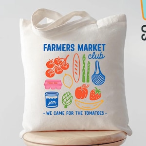 May include: Off-white canvas tote bag with the text "FARMERS MARKET CLUB" and colorful illustrations of produce. The bag features a handle and the phrase "WE CAME FOR THE TOMATOES."