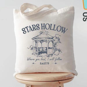 May include: Cream-colored tote bag with a blue gazebo graphic and the words "STARS HOLLOW" above the gazebo. Below the gazebo is the text "Where you lead, I will follow" and "Est.1779". The bag has a light-colored handle and is sitting on a wooden stool.