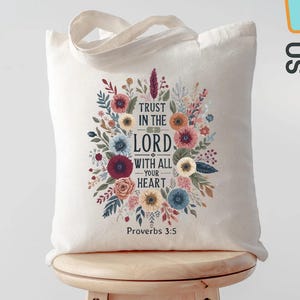 May include: A cream-colored tote bag with a floral design and the text "Trust in the Lord with all your heart" and "Proverbs 3:5". The bag has a simple handle and is sitting on a wooden stool.