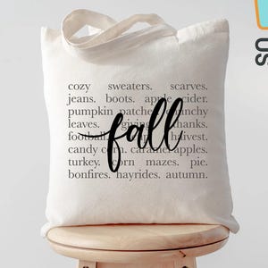 May include: A cream-colored tote bag with the word "fall" in a cursive font. The bag is printed with a list of fall-themed words like "sweaters," "pumpkin patch," and "hayrides." The bag is sitting on a wooden stool.