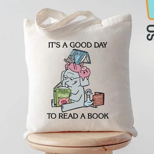 May include: Off-white canvas tote bag featuring a whimsical illustration of an elephant and pig reading books. The text "IT'S A GOOD DAY TO READ A BOOK" is printed above the illustration. The bag has a light beige handle.