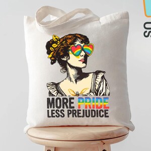 May include: A white tote bag featuring a vintage illustration of a woman wearing rainbow heart-shaped sunglasses. The text "MORE PRIDE LESS PREJUDICE" is printed below the image. The bag has a simple design with two handles.