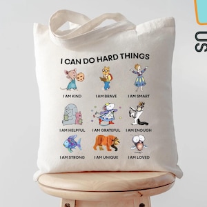 May include: A cream-colored tote bag with the text "I CAN DO HARD THINGS" and illustrations of cartoon characters with positive affirmations. The bag also has the text "I AM KIND, BRAVE, SMART, HELPFUL, GRATEFUL, ENOUGH, STRONG, UNIQUE, LOVED."