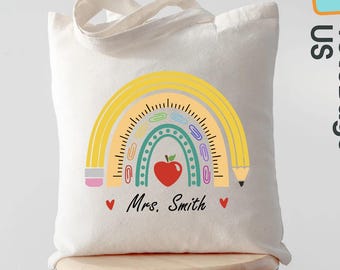 May include: Cream-colored tote bag featuring a colorful rainbow design with a red apple, pencils, and the text "Mrs. Smith." The bag has a teacher theme and is likely used for carrying books or supplies. The bag is sitting on a wooden stool.