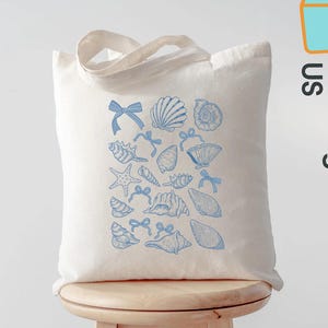 K&ouml;nnte beinhalten: Eine cremefarbene Tragetasche mit einem blauen Muschel- und Schleifenmuster. Die Tasche hat eine rechteckige Form und zwei Henkel. Der Text "Custom ToteBags US" ist auf der Seite aufgedruckt.