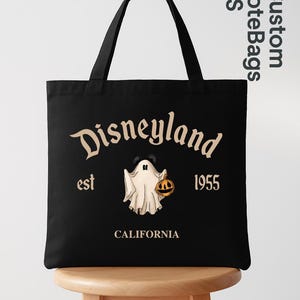 May include: Black tote bag featuring the Disneyland logo with a ghost holding a jack-o'-lantern. The text "Disneyland est 1955 California" is printed in a vintage style. The bag has black handles and is displayed on a wooden stool.
