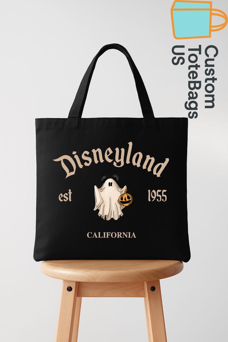 May include: Black tote bag featuring the Disneyland logo with a ghost holding a jack-o'-lantern. The text "Disneyland est 1955 California" is printed in a vintage style. The bag has black handles and is displayed on a wooden stool.