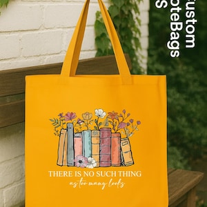 May include: Mustard yellow tote bag with a graphic of books and flowers. The text "THERE IS NO SUCH THING as too many books" is printed below the graphic. The bag has long handles and is sitting on a wooden bench.