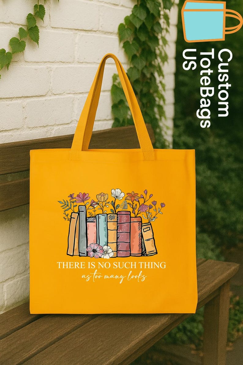 May include: Mustard yellow tote bag with a graphic of books and flowers. The text "THERE IS NO SUCH THING as too many books" is printed below the graphic. The bag has long handles and is sitting on a wooden bench.
