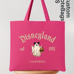 May include: A pink tote bag featuring the Disneyland logo with a ghost holding a pumpkin. The text "est 1955 CALIFORNIA" is also displayed. The bag has two pink handles and is sitting on a wooden stool.