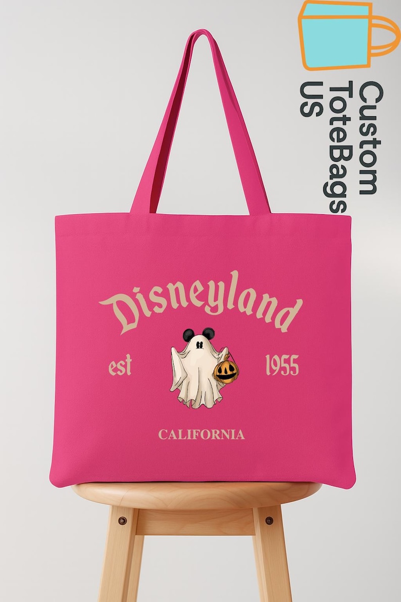 May include: A pink tote bag featuring the Disneyland logo with a ghost holding a pumpkin. The text "est 1955 CALIFORNIA" is also displayed. The bag has two pink handles and is sitting on a wooden stool.